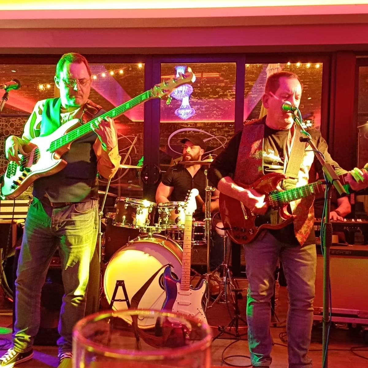 A band performing on stage with a bassist, drummer, and guitarist. The stage is lit with pink and yellow lights. A glass is visible in the foreground.