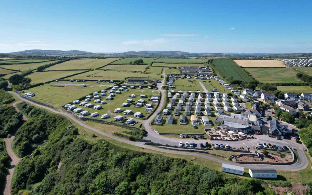 Aerial view of a campground with numerous caravans arranged in rows on open green fields. Surrounding area features hedged plots, a road, and a few scattered buildings under a clear blue sky.
