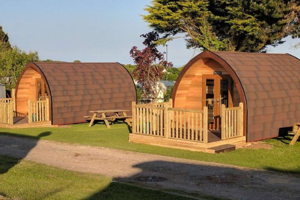Two wooden glamping pods with curved roofs sit on grass. Each has a small porch and picnic table. Tall trees and a clear sky are in the background.