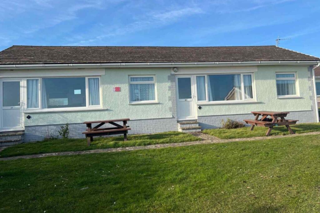 Single-story holiday bungalow with light-coloured brick exterior, two doors and windows, and two picnic tables on grass in front. Blue sky overhead.