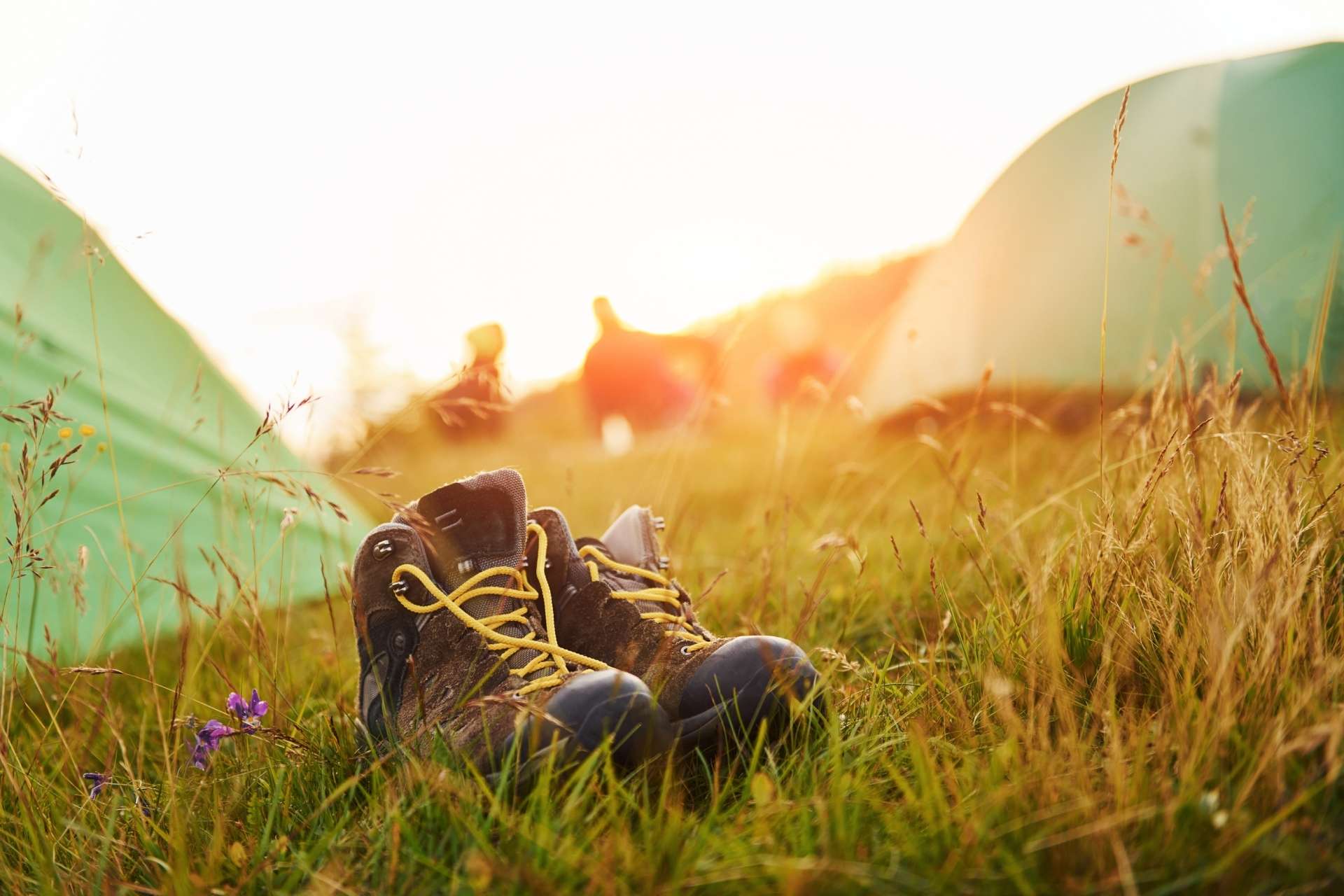 A pair of hiking boots with yellow laces rests on grass in a sunlit campsite, with blurred tents and people in the background.