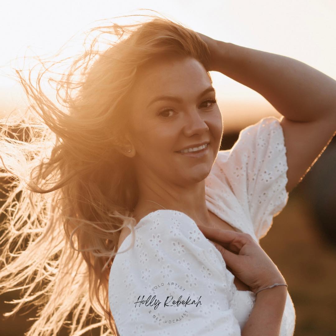 A person with long hair wearing a white top, smiling and holding their hair in a bright outdoor setting with sunlight in the background.