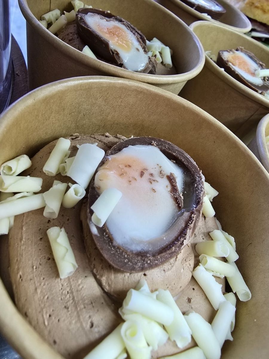 A chocolate dessert topped with a halved chocolate shell resembling a soft-boiled egg and white chocolate curls, served in a brown paper bowl.