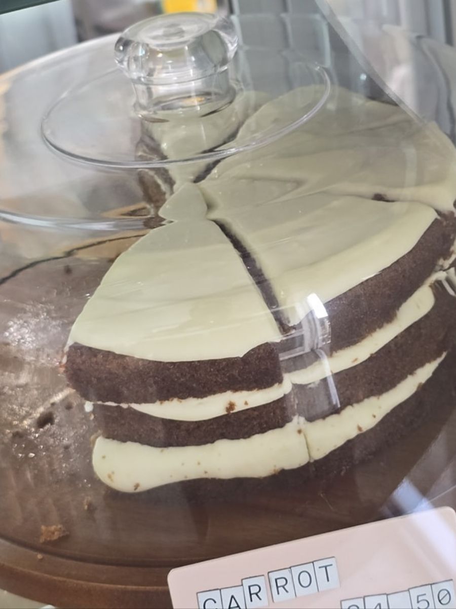 A three-layer carrot cake with white frosting, partially sliced, displayed under a glass dome.