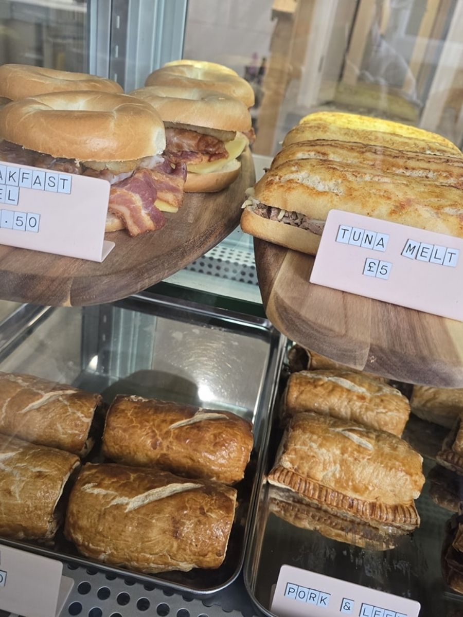 Display case with breakfast bagels, tuna melt sandwiches, sausage rolls, and pasties, each on wooden boards with price labels.