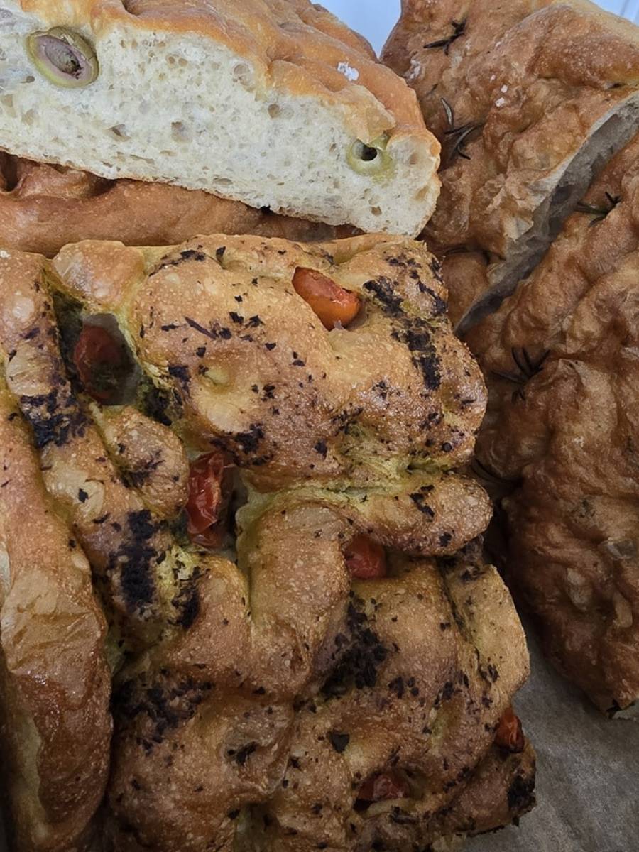 Several loaves of focaccia bread with different toppings, including olives and tomatoes, are stacked closely together.