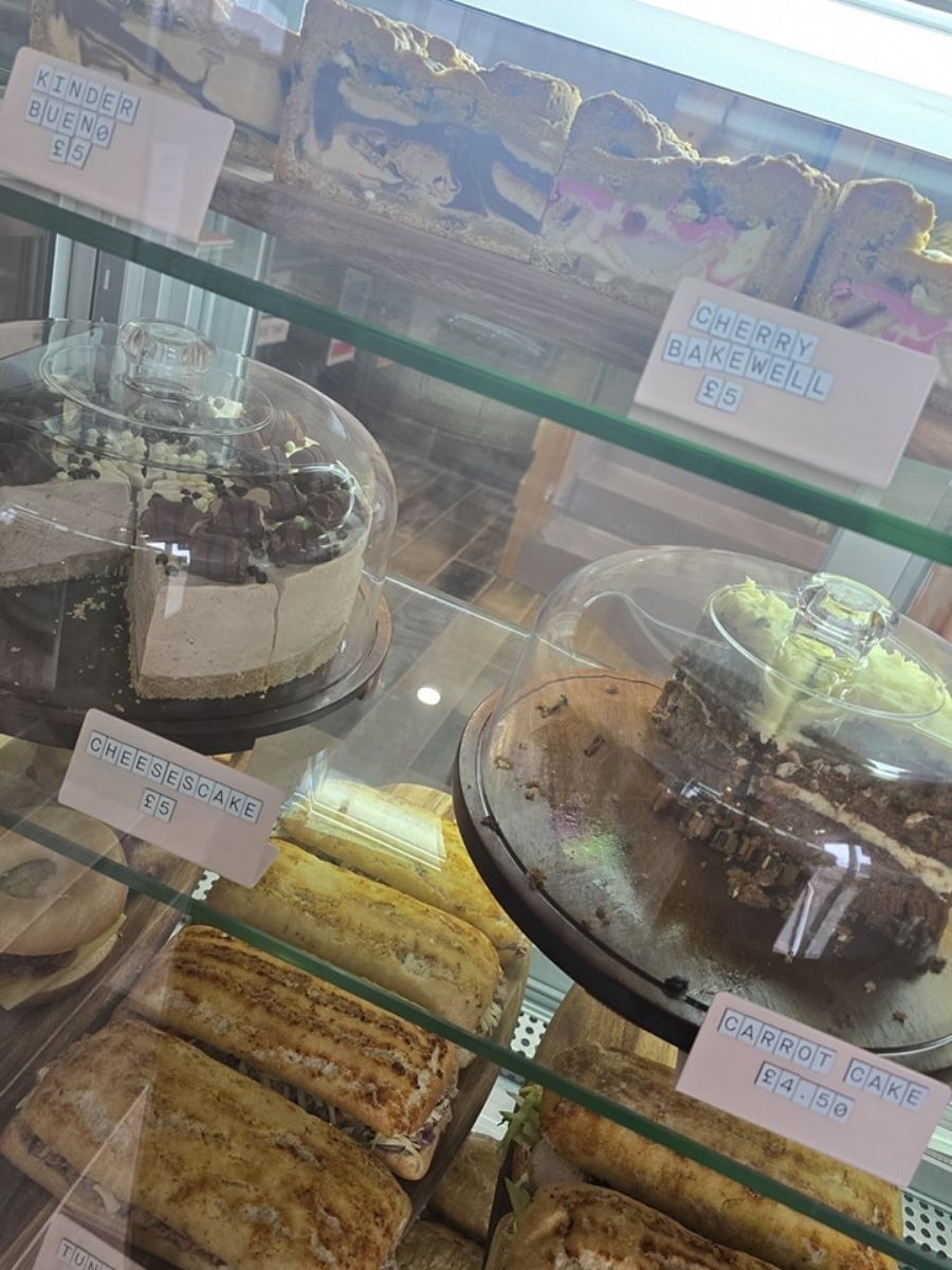 A bakery display case contains cakes, slices, and pastries under glass domes and on shelves, with labeled signs for each item and prices visible.