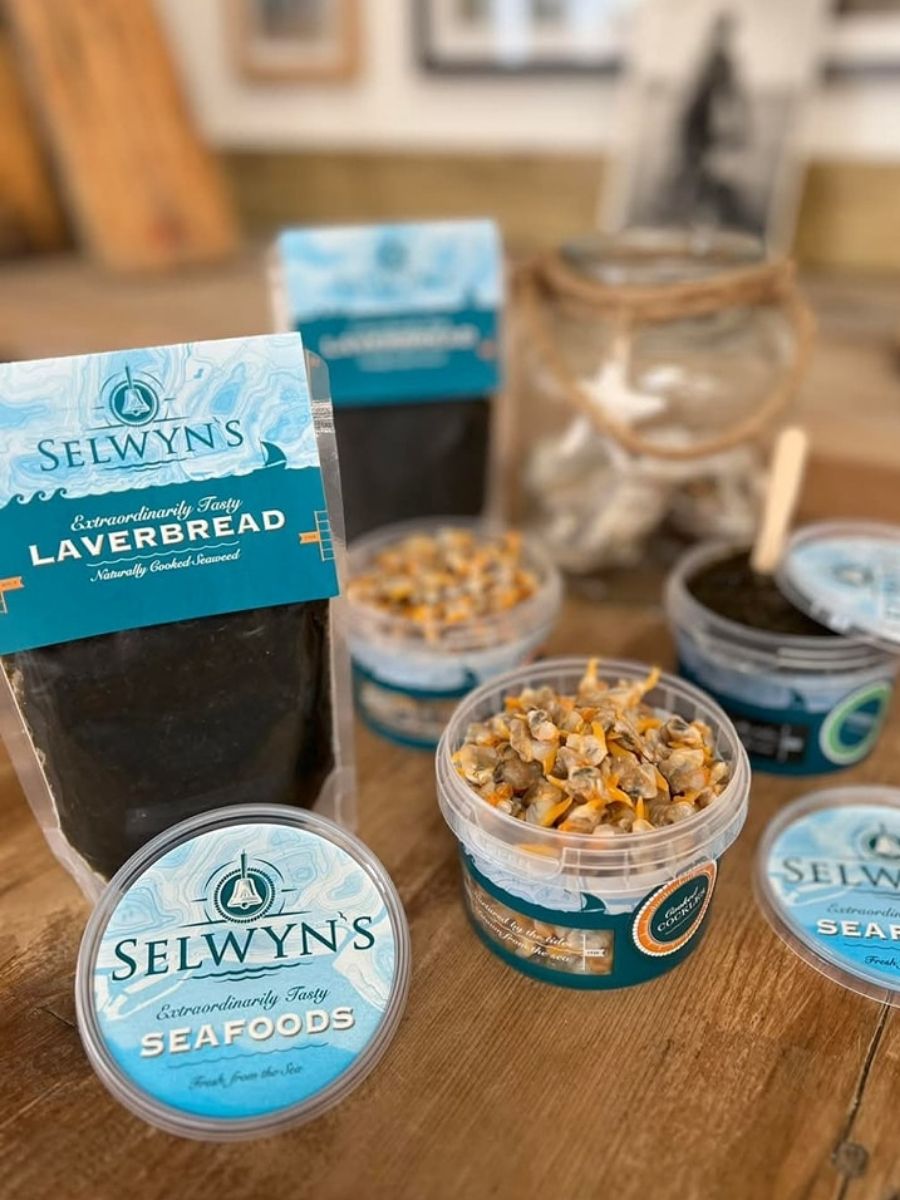 Assorted Selwyn’s branded seafood products, including laverbread and seafood cups, displayed on a wooden surface with a glass jar in the background.