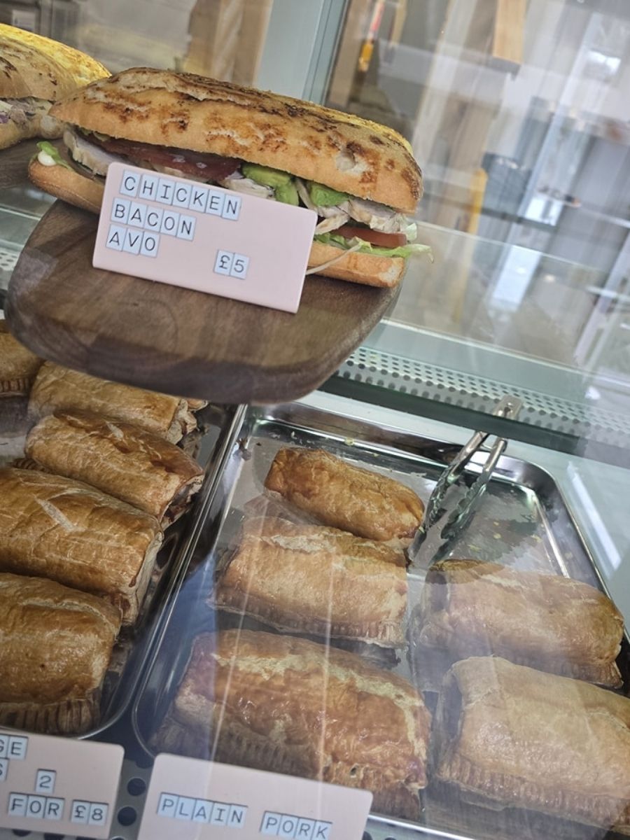 A chicken, bacon, and avocado sandwich is displayed above trays of sausage rolls and pastries in a glass bakery case.