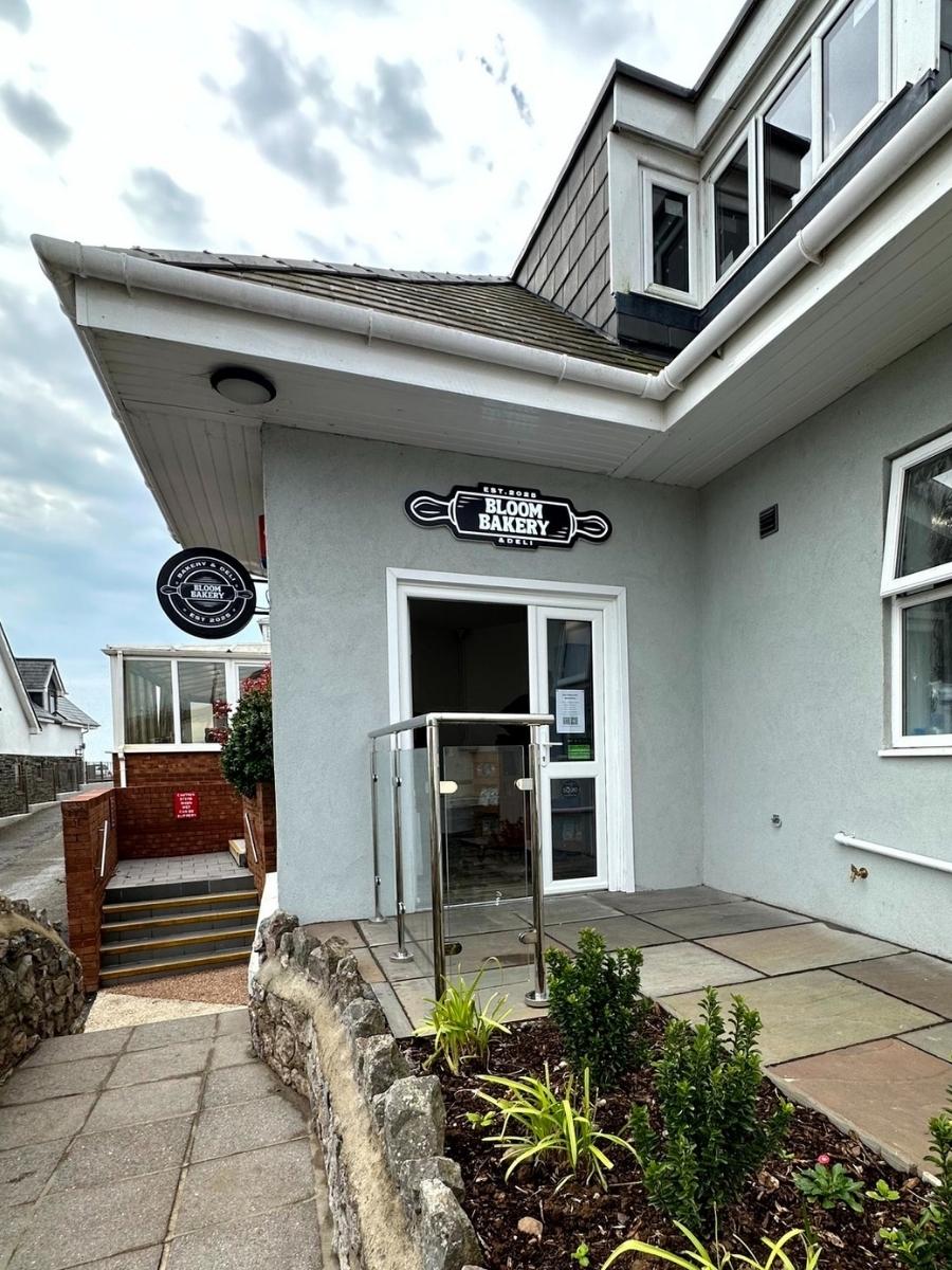 Entrance to Bloom Bakery, a grey building with a sign above the door and a glass railing, located on a stone path with small plants in front.
