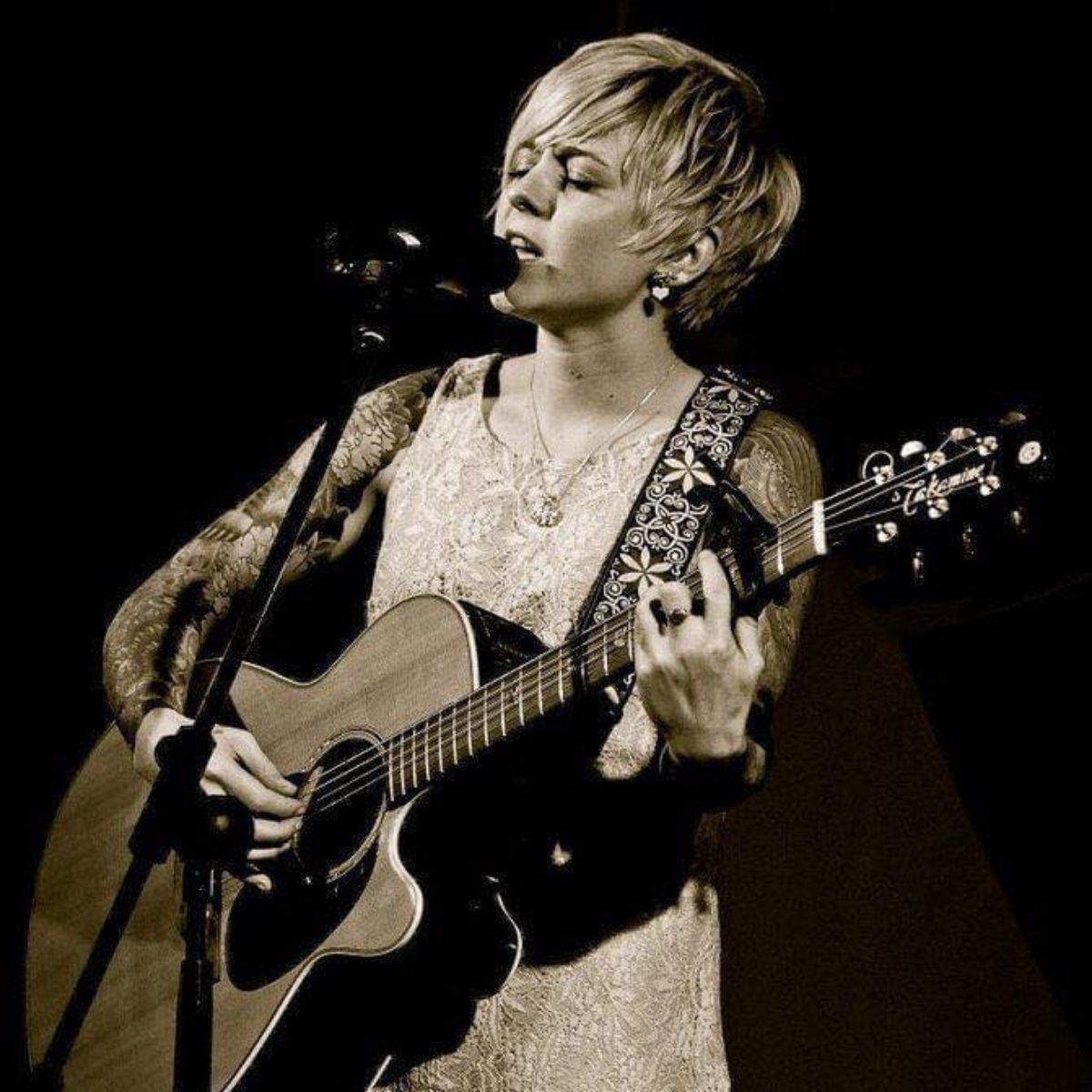 A person with short hair and tattoos plays an acoustic guitar and sings into a microphone on stage.