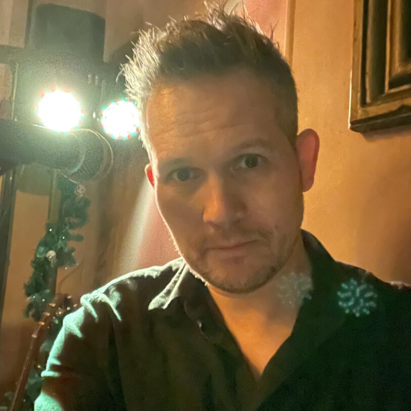 A man with short hair stands indoors near a microphone and bright stage lights, looking directly at the camera.