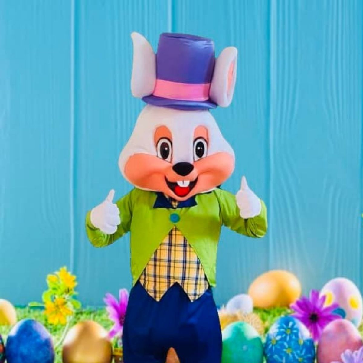 Person in an Easter Bunny costume with a purple top hat, green jacket, and plaid vest stands in front of decorated eggs and flowers against a blue background.