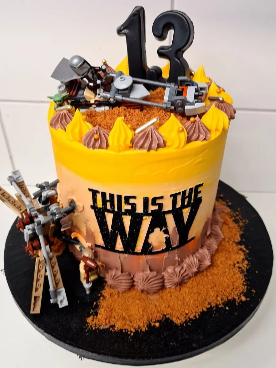 A round cake with yellow and brown frosting, a black "13" topper, LEGO Star Wars figures, and "THIS IS THE WAY" text on the side.