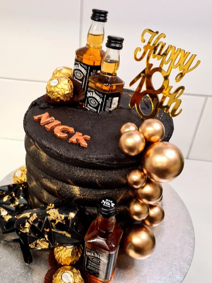A black and gold birthday cake decorated with Ferrero Rocher chocolates, gold balloons, and Jack Daniel’s miniature bottles. The cake topper reads "Happy 18th" and "Nick".