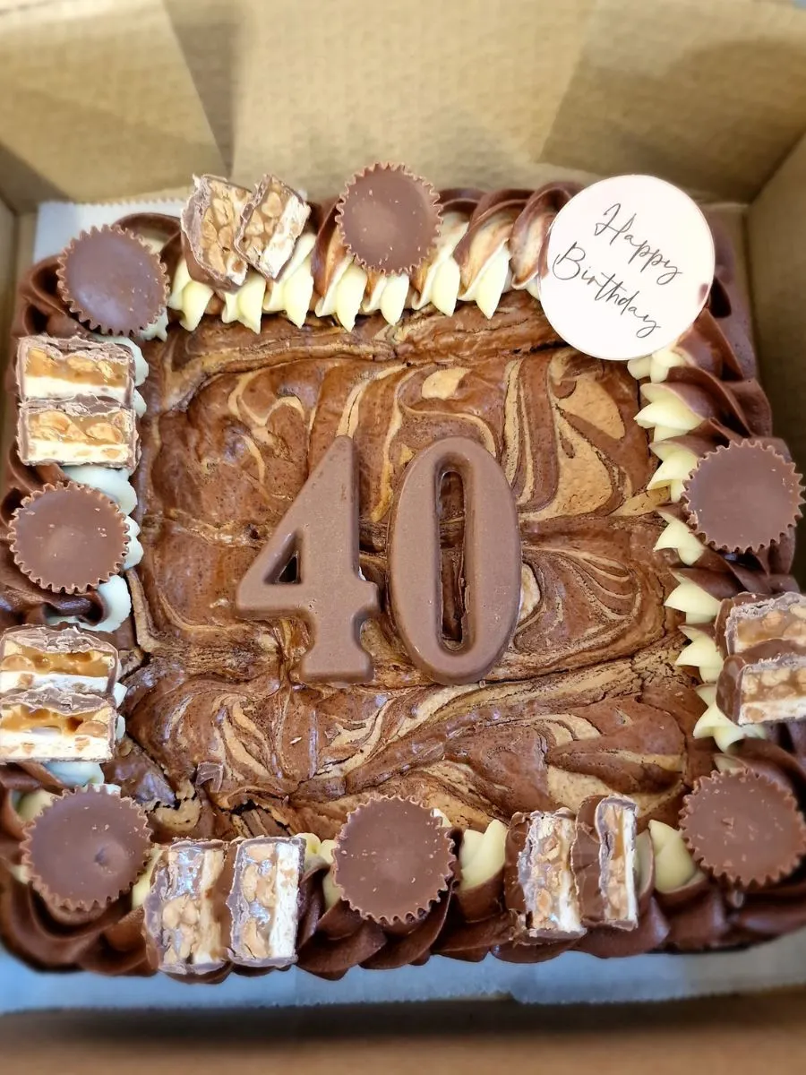 A square chocolate cake with "40" in chocolate on top, decorated with peanut butter cups, snickers pieces, and a white disc that says "Happy Birthday".