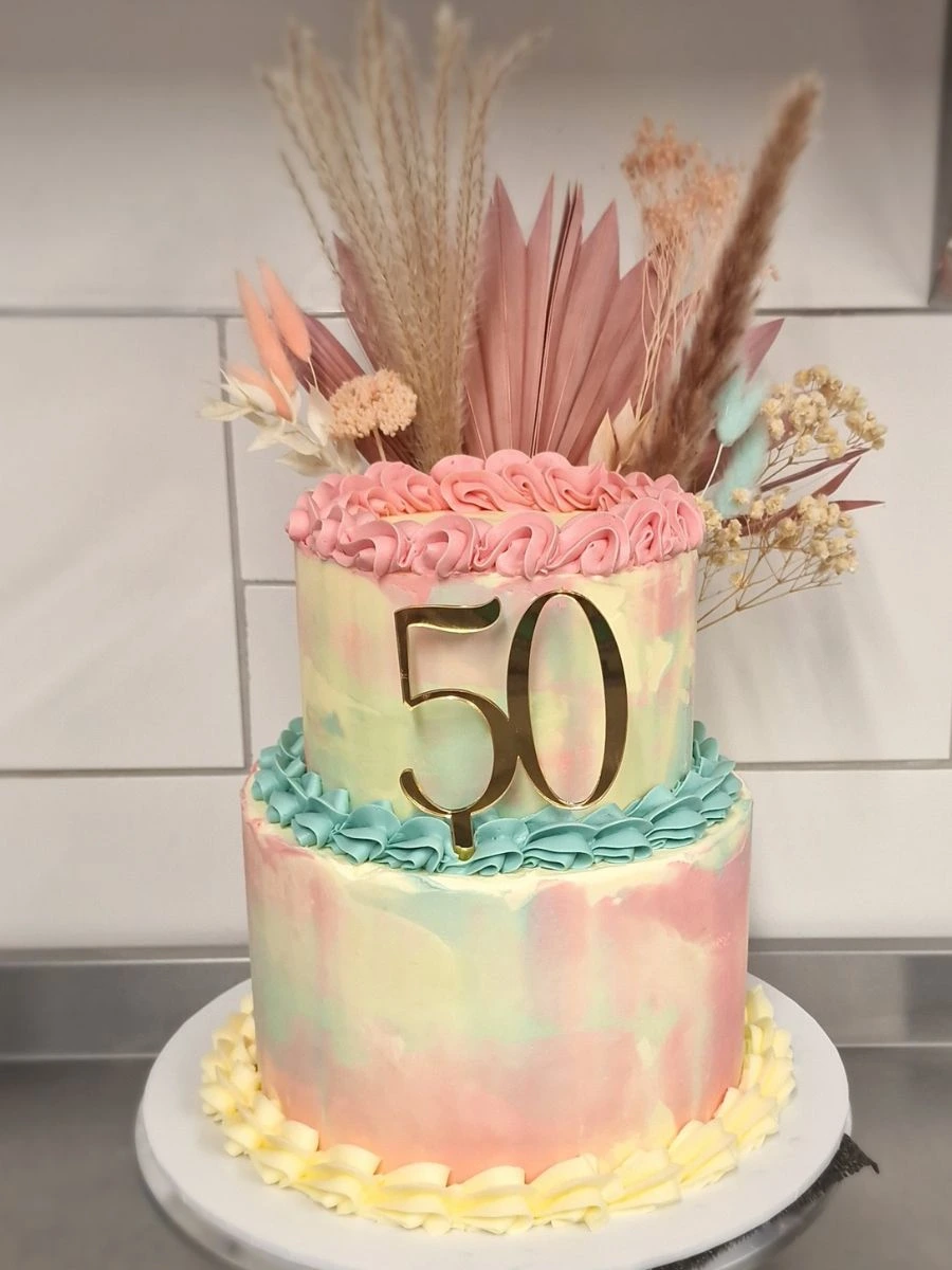 Two-tier pastel cake decorated with buttercream ruffles, a gold "50" topper, and dried floral arrangements on top.