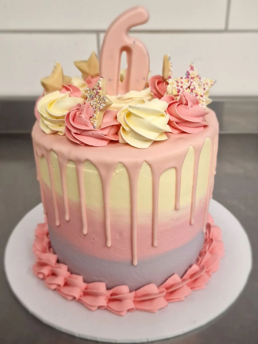 A pastel pink, purple, and yellow drip cake decorated with rosettes, star-shaped toppers, and a large number 6 on top, placed on a white cake board.