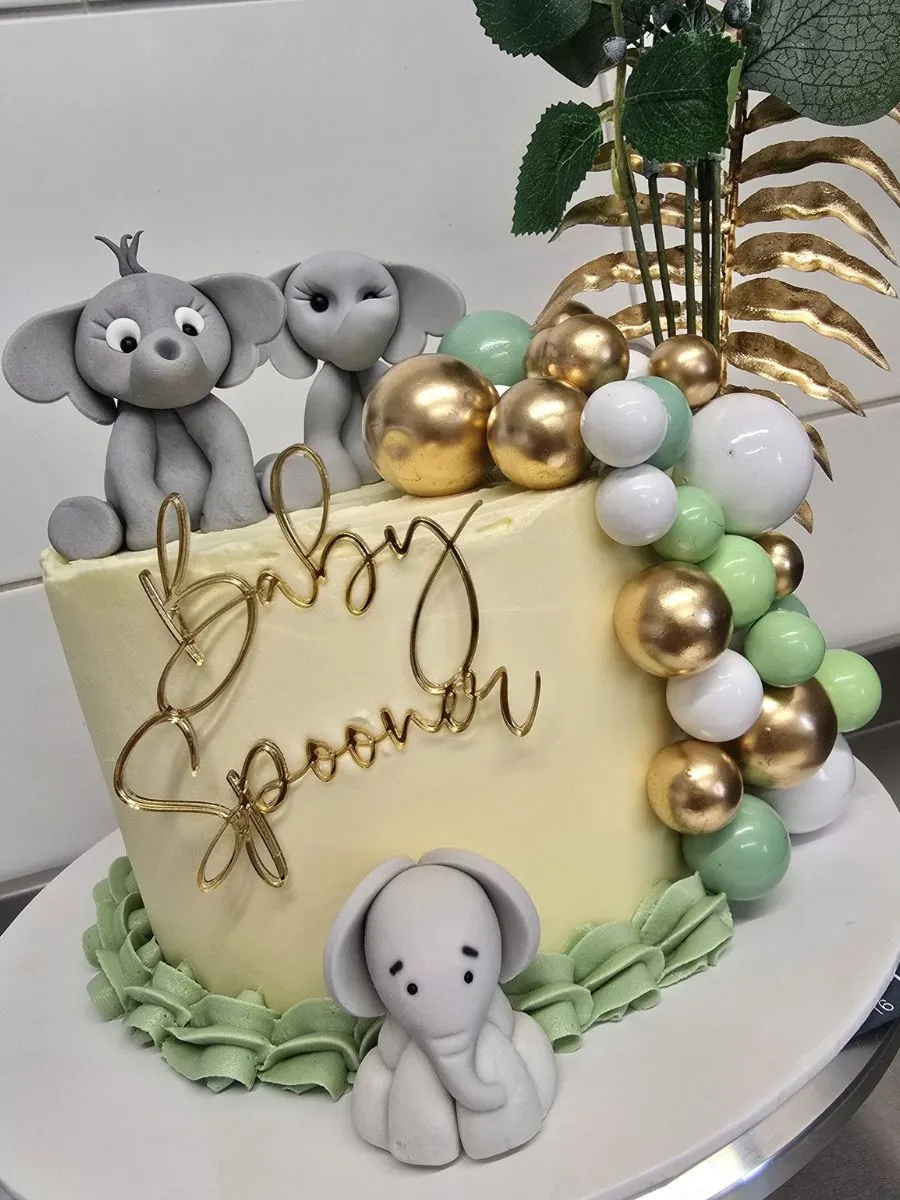 A round cake decorated with fondant elephant figurines, gold and pastel balloons, green ruffles, and a topper reading "Baby Spooner." Green leaves are arranged at the back.