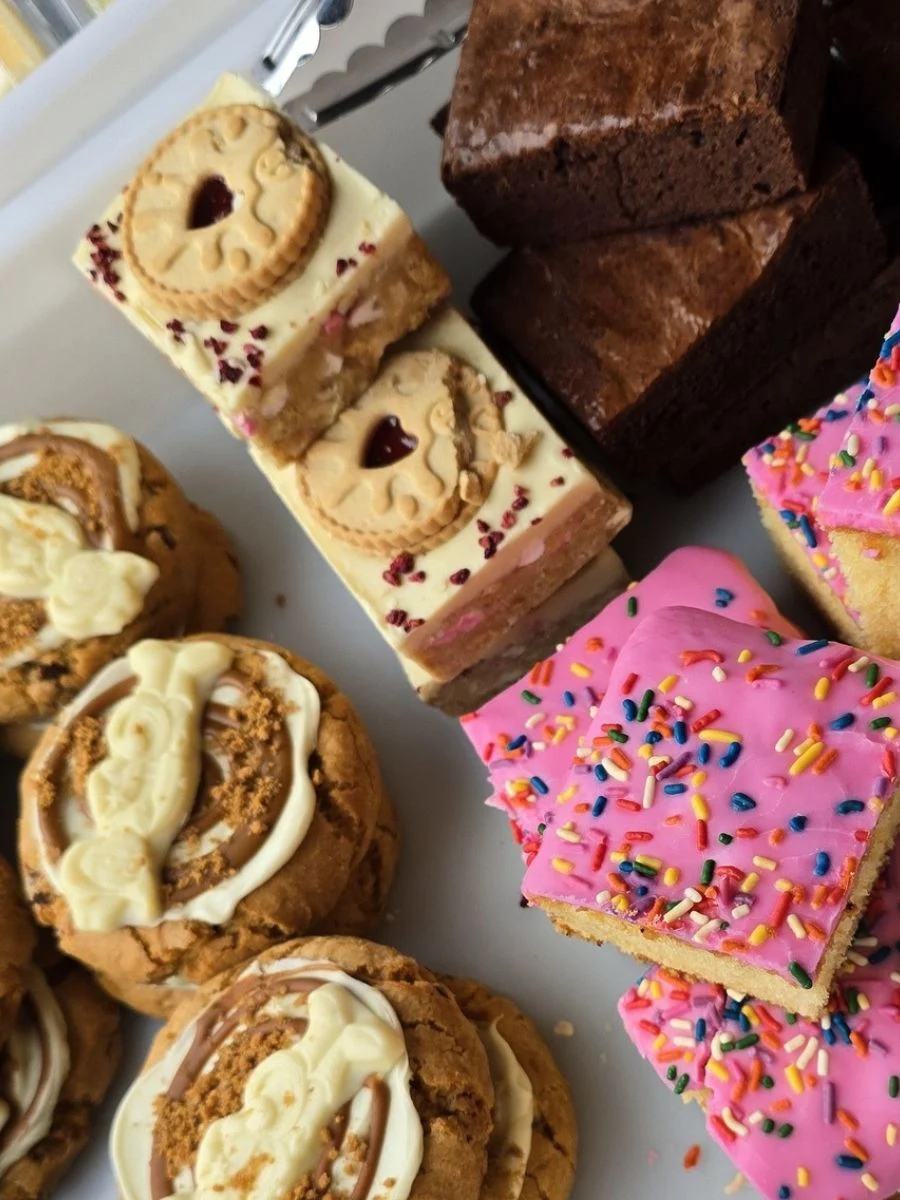 A tray with cookies topped with white chocolate, layered biscuits with jam, chocolate brownies, and pink-iced cake squares with sprinkles.