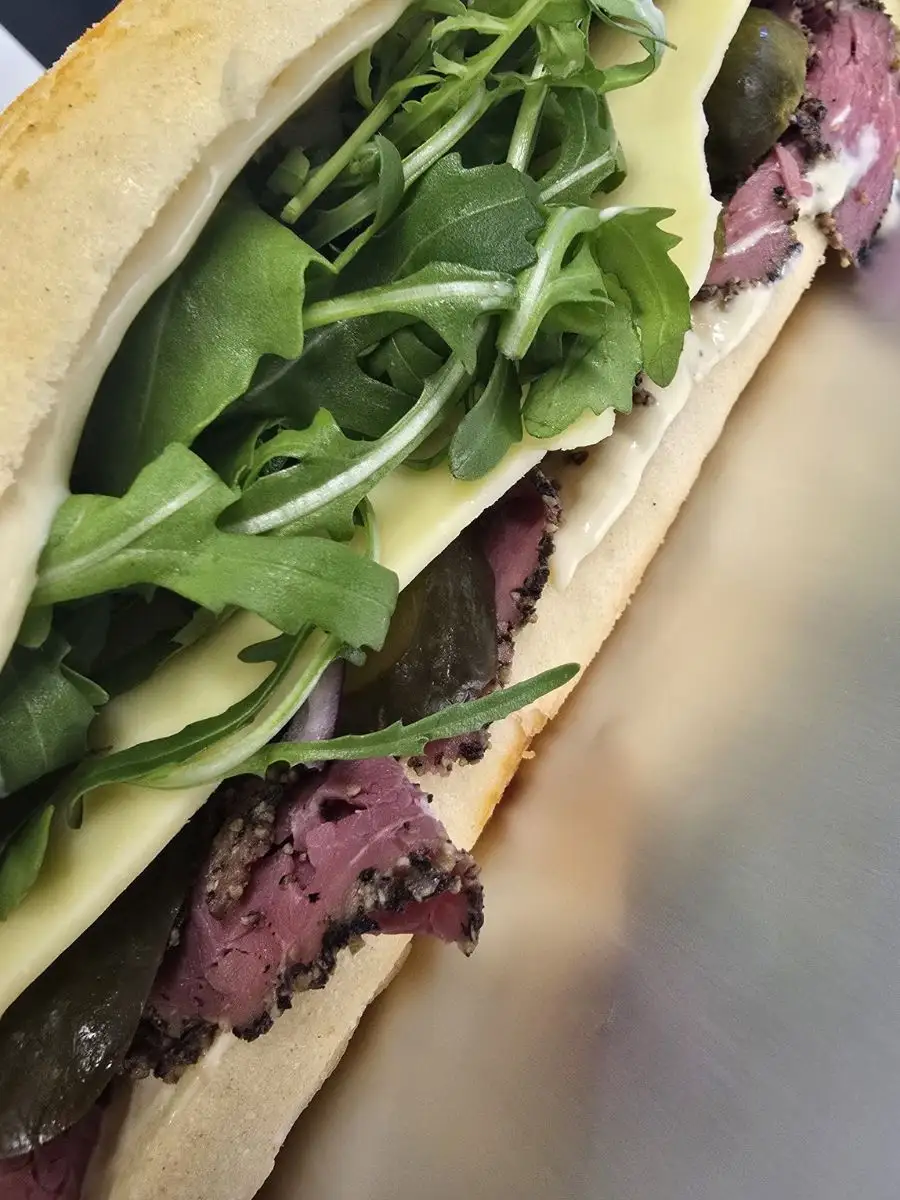 A close-up of a sandwich filled with slices of seasoned meat, rocket, cheese, and leafy greens on a white sub roll.