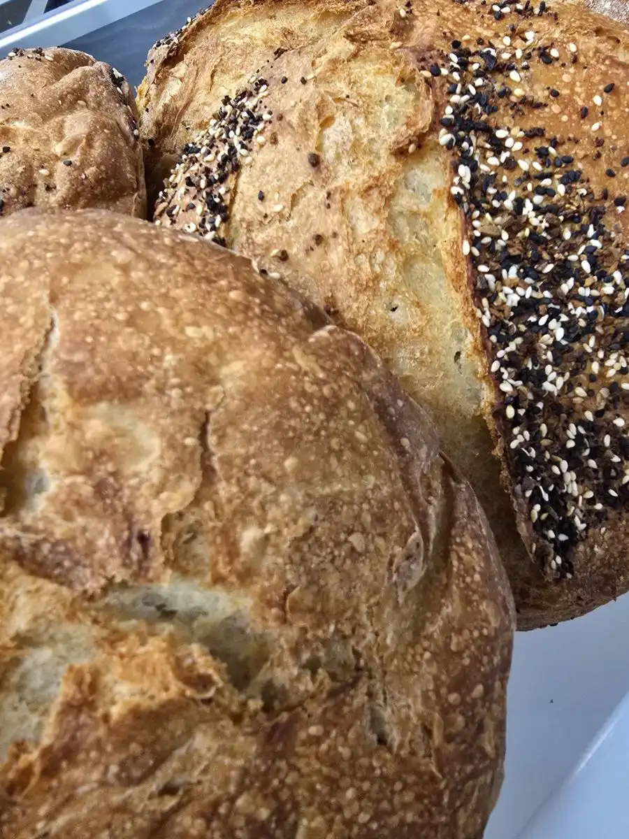Two round loaves of artisan bread on a tray, one topped with mixed seeds, both featuring a golden, crusty exterior.