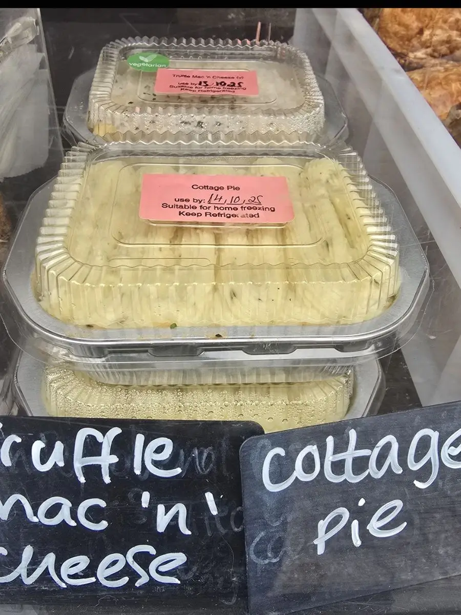 Clear plastic containers of cottage pie are displayed in a refrigerated case, each with a printed label on top, next to a chalkboard sign reading “cottage pie.”.